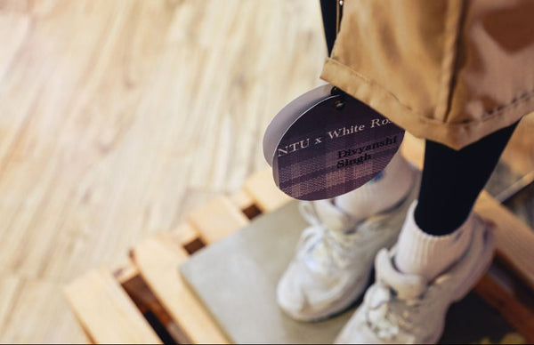 mannequin wearing white sneakers and a beige jacket with a product container labeled 'NTU x White Rose' on a wooden floor.